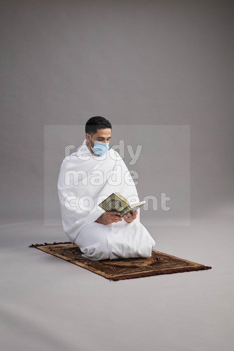 A man wearing Ehram with face mask sitting on floor reading quran on gray background