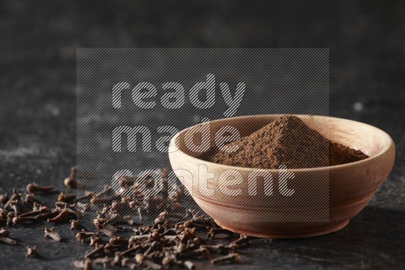 A wooden bowl full of cloves powder on a textured black flooring