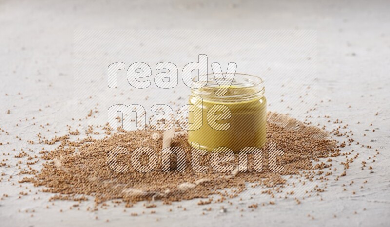 A glass jar full of mustard paste set on a burlap piece with a wooden spoon full of mustard seeds on a textured white flooring