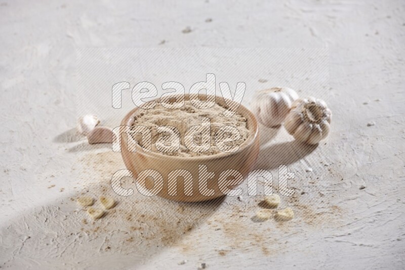 A wooden bowl full of garlic powder and beside it garlic cloves on a textured white flooring in different angles