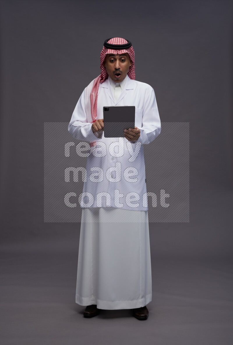 Saudi man wearing thob with lab coat and shomag with pocket employee badge standing working on tablet on gray background