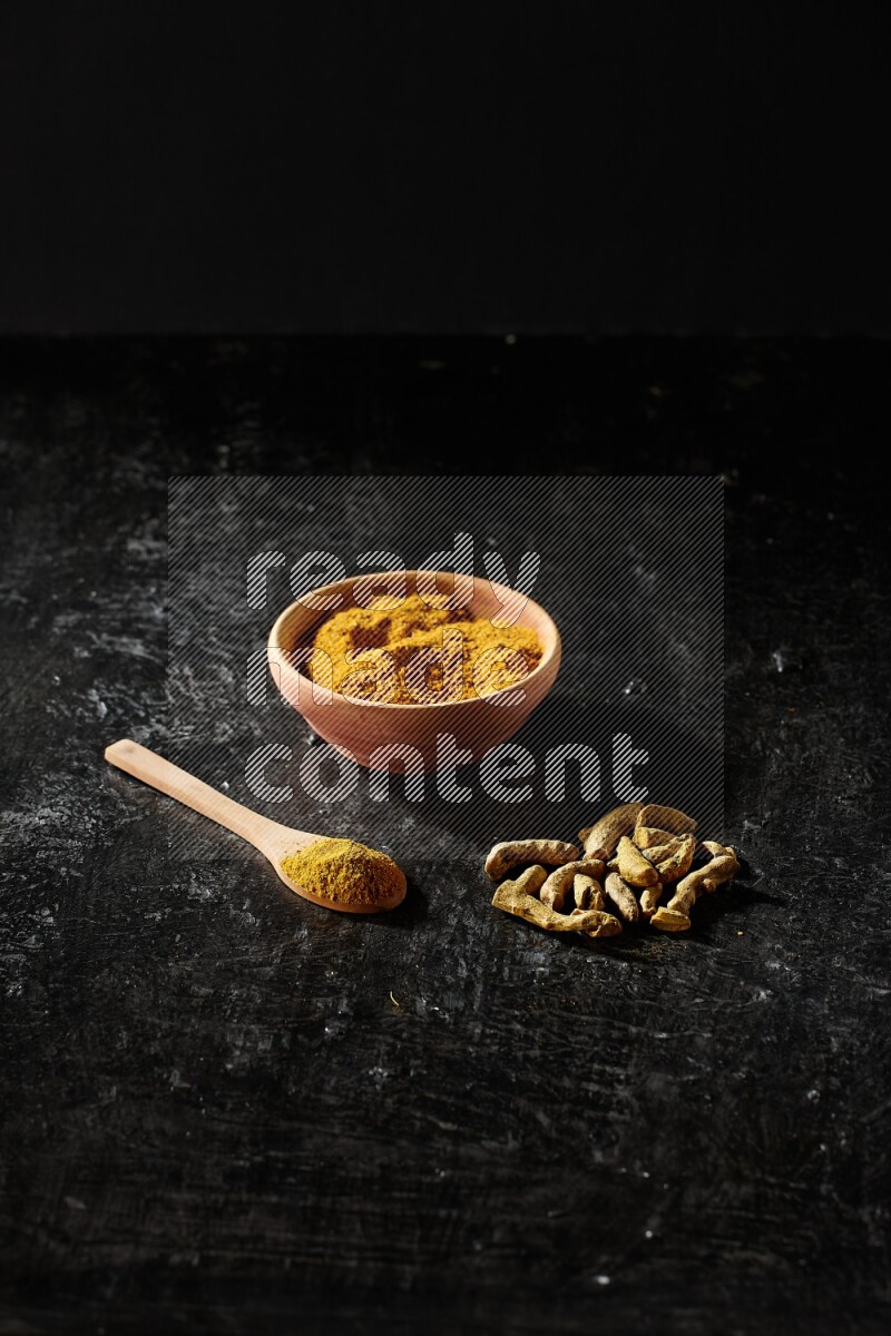 A wooden bowl and a wooden spoon full of turmeric powder with dried turmeric fingers on textured black flooring