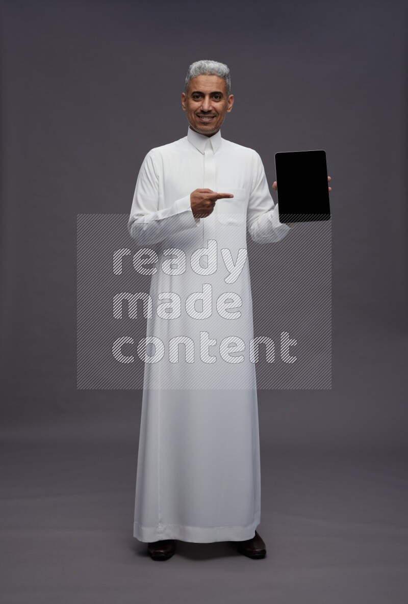 Saudi man wearing thob standing showing tablet to camera on gray background