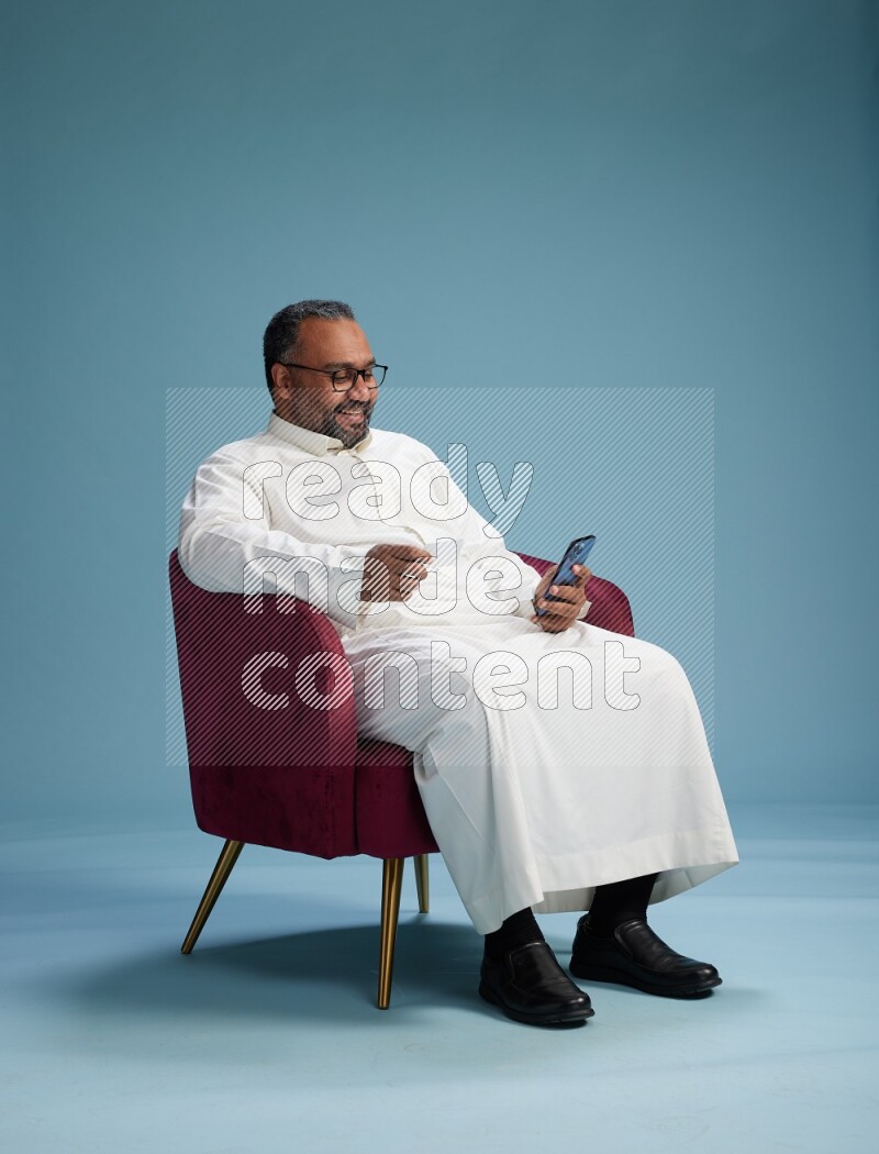 Saudi Man without shimag sitting on chair holding ATM while talking on phone on blue background