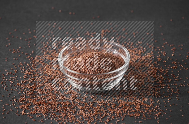 A glass bowl full of garden cress seeds surrounded by seeds on a black flooring