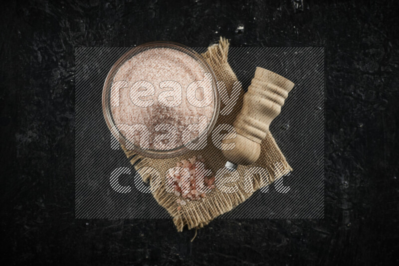 A glass bowl full of pink himalayan salt with a wooden grinder on a burlap fabric all on black background