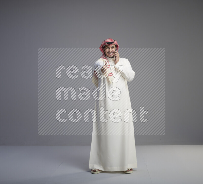 A Saudi man standing wearing thob and red shomag talking on phone on gray background