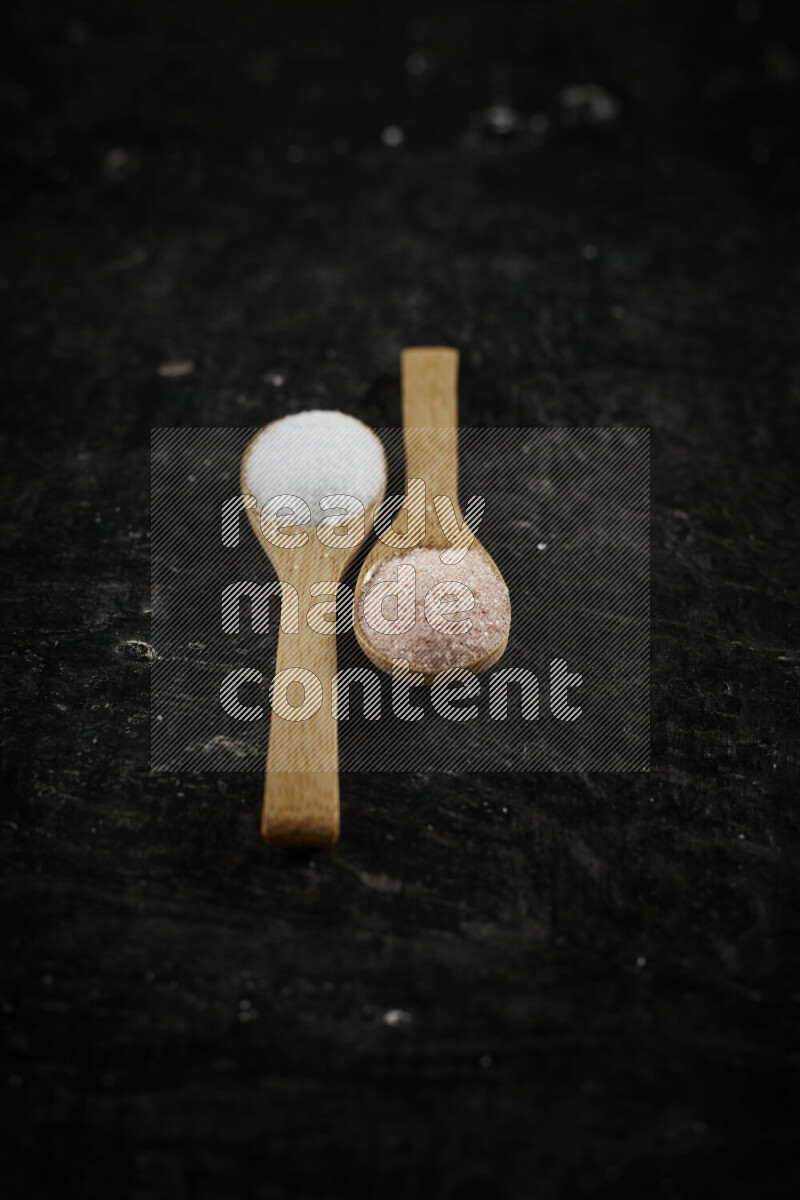 2 wooden spoons filled with fine and coarse salt on black background