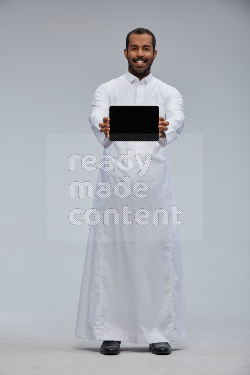 Saudi man wearing Thob standing showing tablet to camera on Gray background