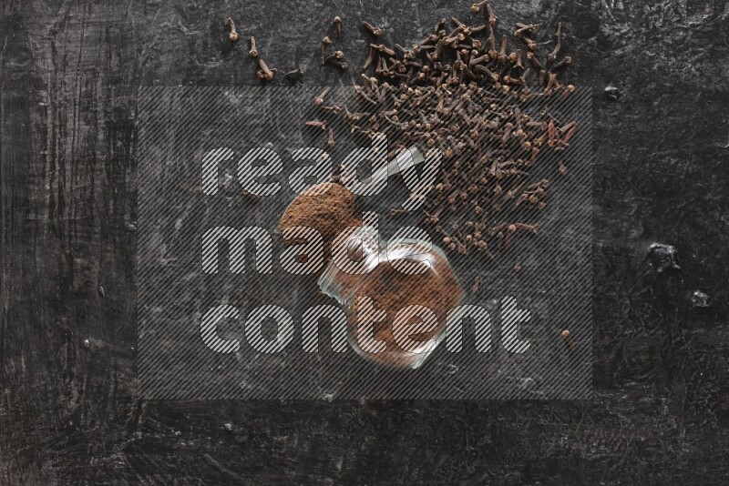 A flipped glass spice jar and a metal spoon full of cloves powder with cloves spread on textured black flooring