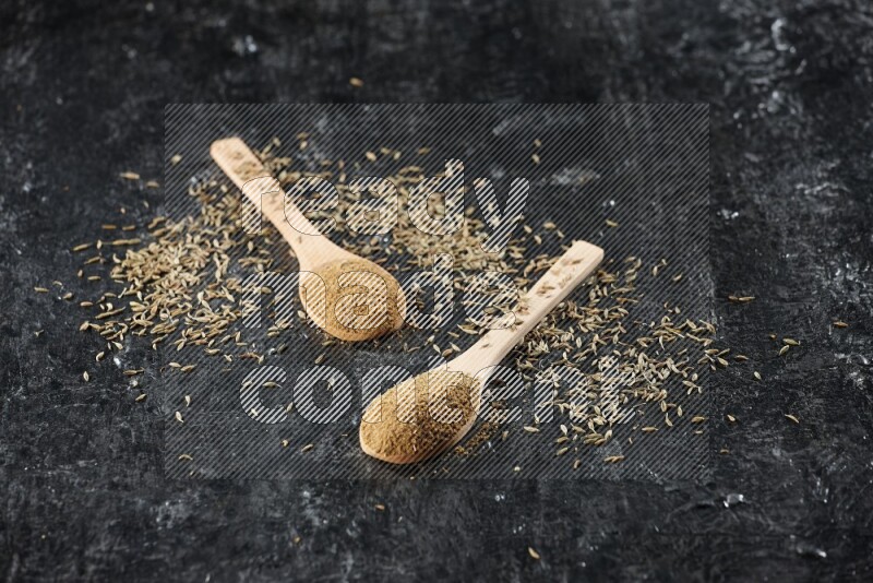 2 wooden spoons full of cumin powder with spreaded seeds on textured black flooring
