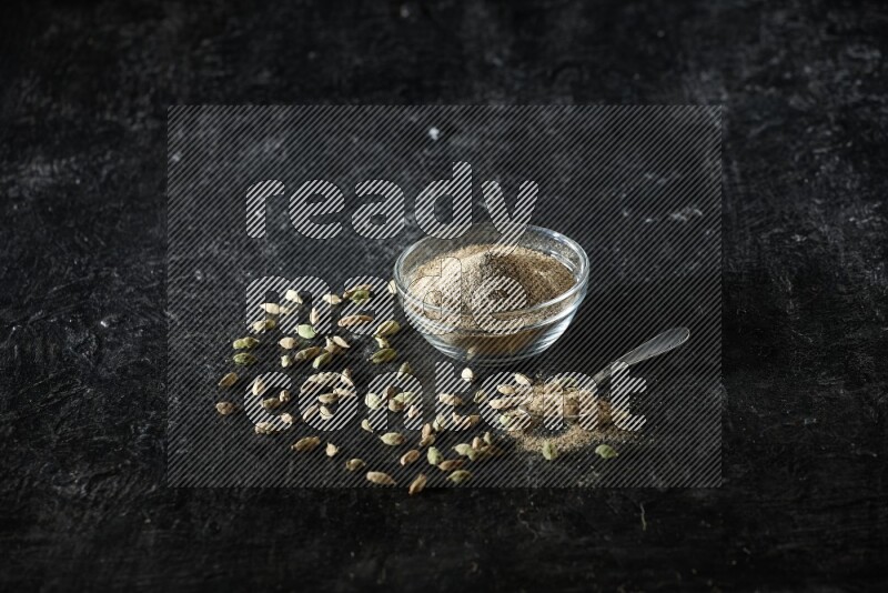 A glass bowl and a metal spoon full of cardamom powder with cardamom seeds beside them on textured black flooring