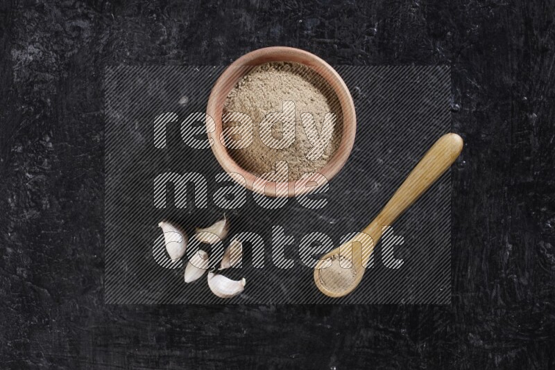 A wooden bowl and spoon full of garlic powder and beside it garlic cloves on a textured black flooring