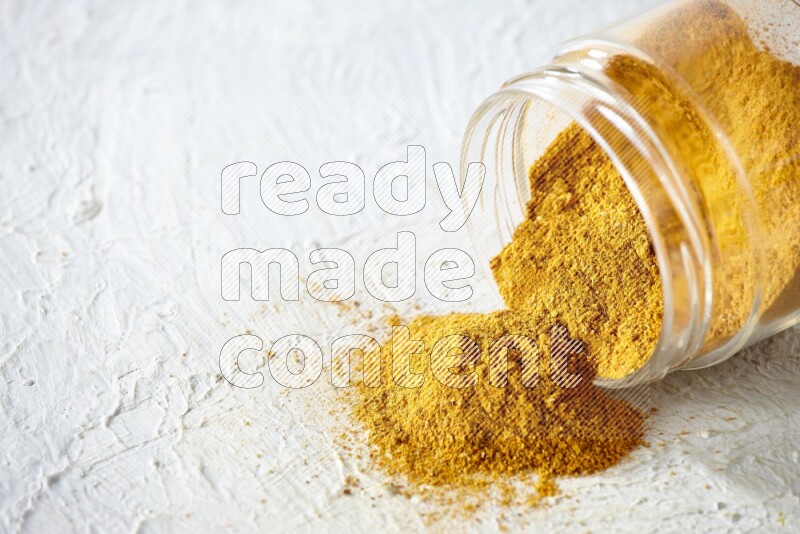 A flipped glass spice jar full of turmeric powder and powder fell out it on textured white flooring