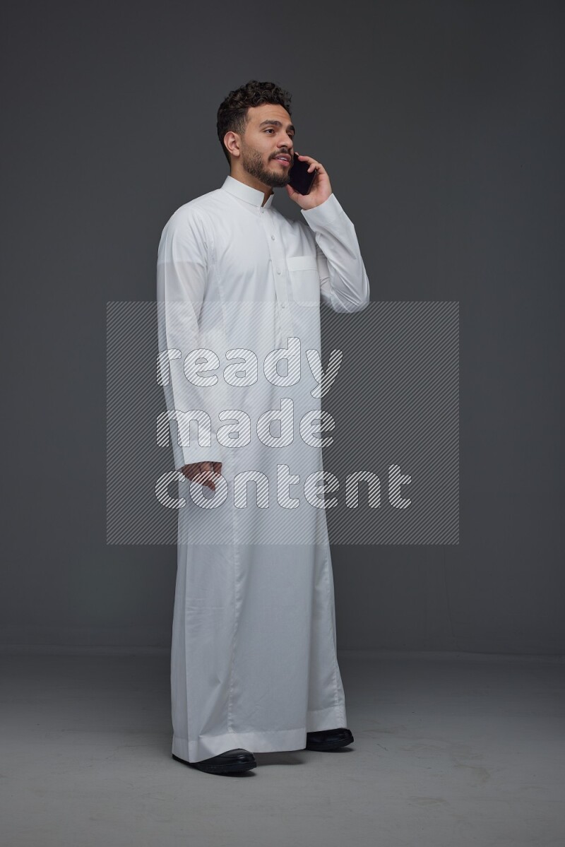 A Saudi man wearing Thobe and talking in the phone while standing and making different poses eye level on a gray background