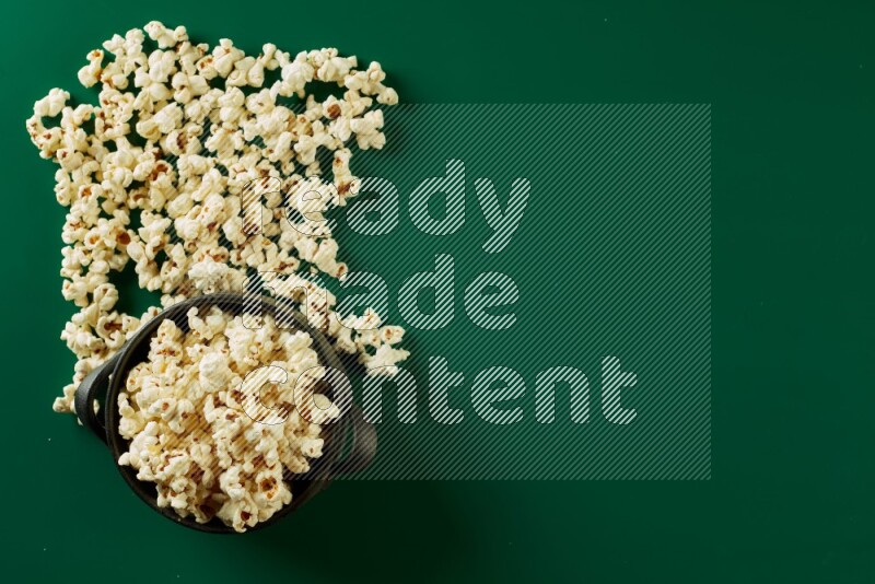 A black ceramic bowl full of popcorn with popcorn beside it on a green background in a top view shot