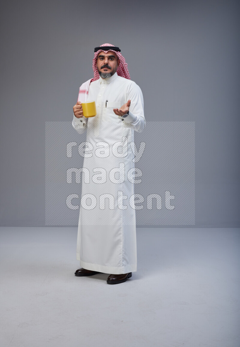 Saudi man Wearing Thob and red Shomag standing holding mug on Gray background