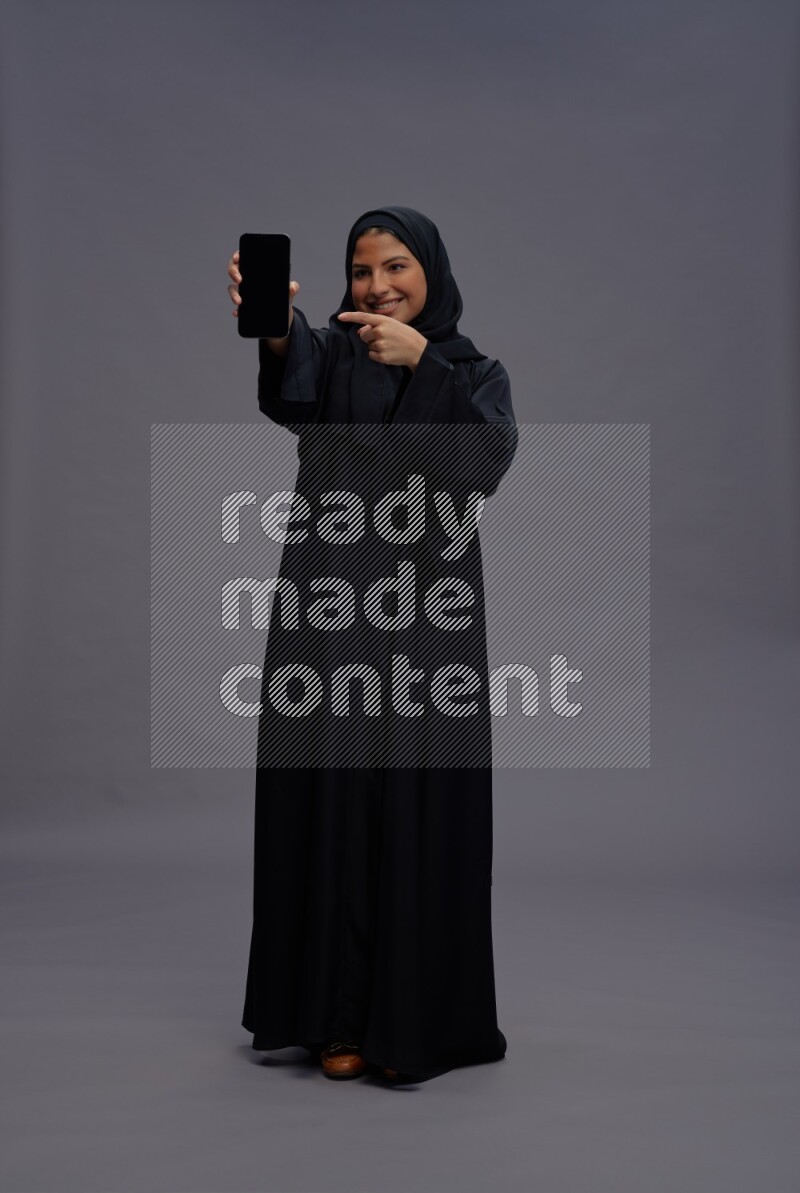 Saudi woman wearing Abaya standing showing phone to camera on gray background