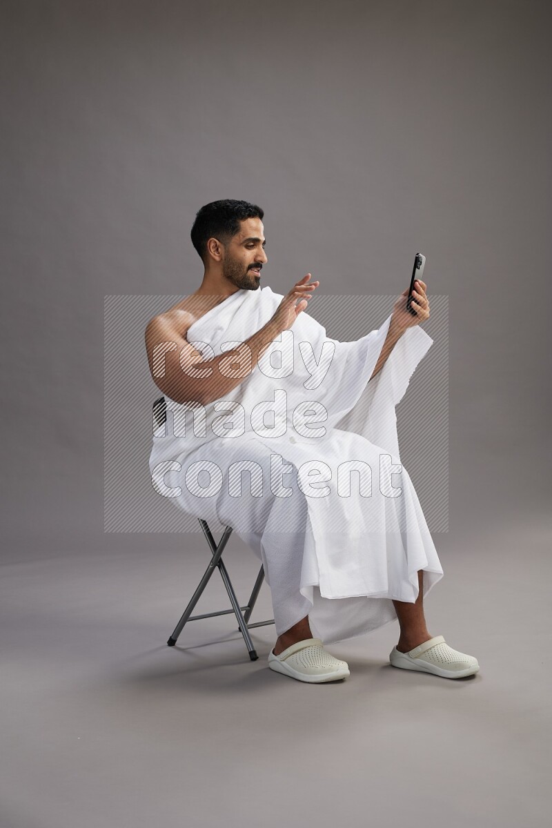 A man wearing Ehram sitting on chair taking selfie on gray background