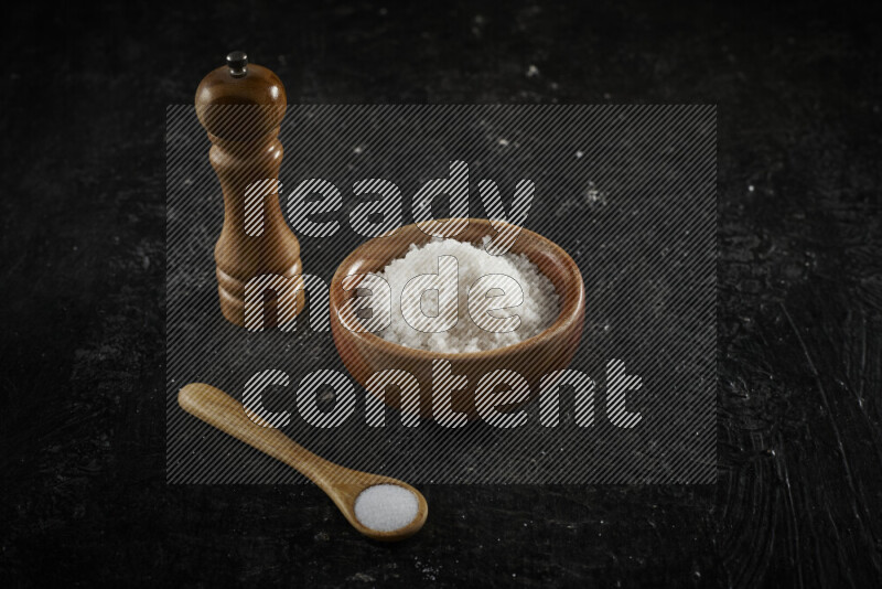 A wooden bowl and spoon filled with white sea salt and wooden grinder beside them on black background