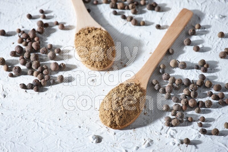 Wooden spoons full of allspice powder and allspice whole balls spreaded on a textured white flooring