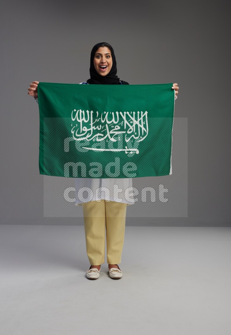Saudi woman wearing lab coat with stethoscope standing holding Saudi flag on Gray background