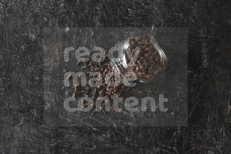 وعاء زجاجي للبهارات مقلوب ممتلئ بالقرنفل والقرنفل سقط منه على أرضية سوداء