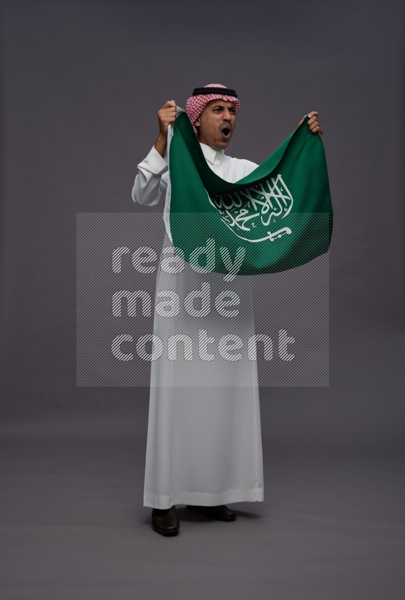Saudi man wearing thob and shomag standing holding Saudi flag on gray background
