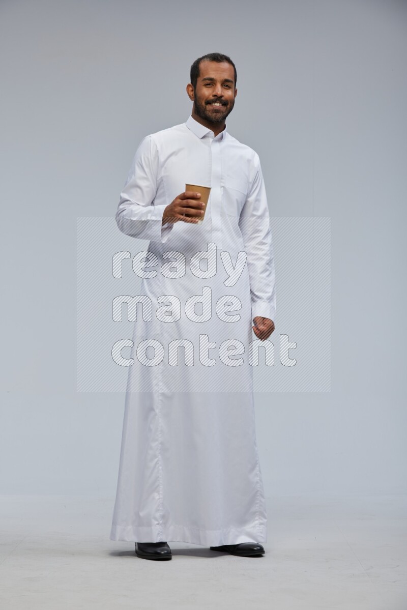 Saudi man wearing thob standing holding paper cup on gray background