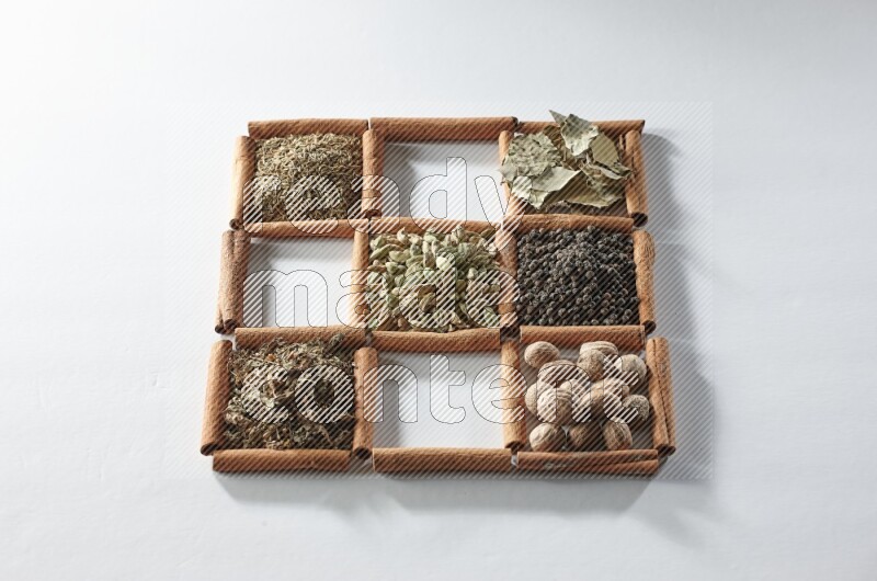 9 مربعات من أعواد القرفة ممتلئة بالهيل في المنتصف محاطة بجوزة الطيب والملح والريحان المجفف والفلفل الأبيض والكمون وبهار الفلفل الحلو وأوراق الغار والفلفل الأسود على أرضية بيضاء