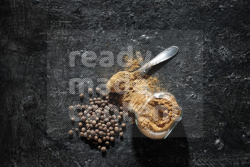 A flipped glass spice jar and metal spoon full of allspice powder and powder spilled out of it with whole balls on a textured black flooring