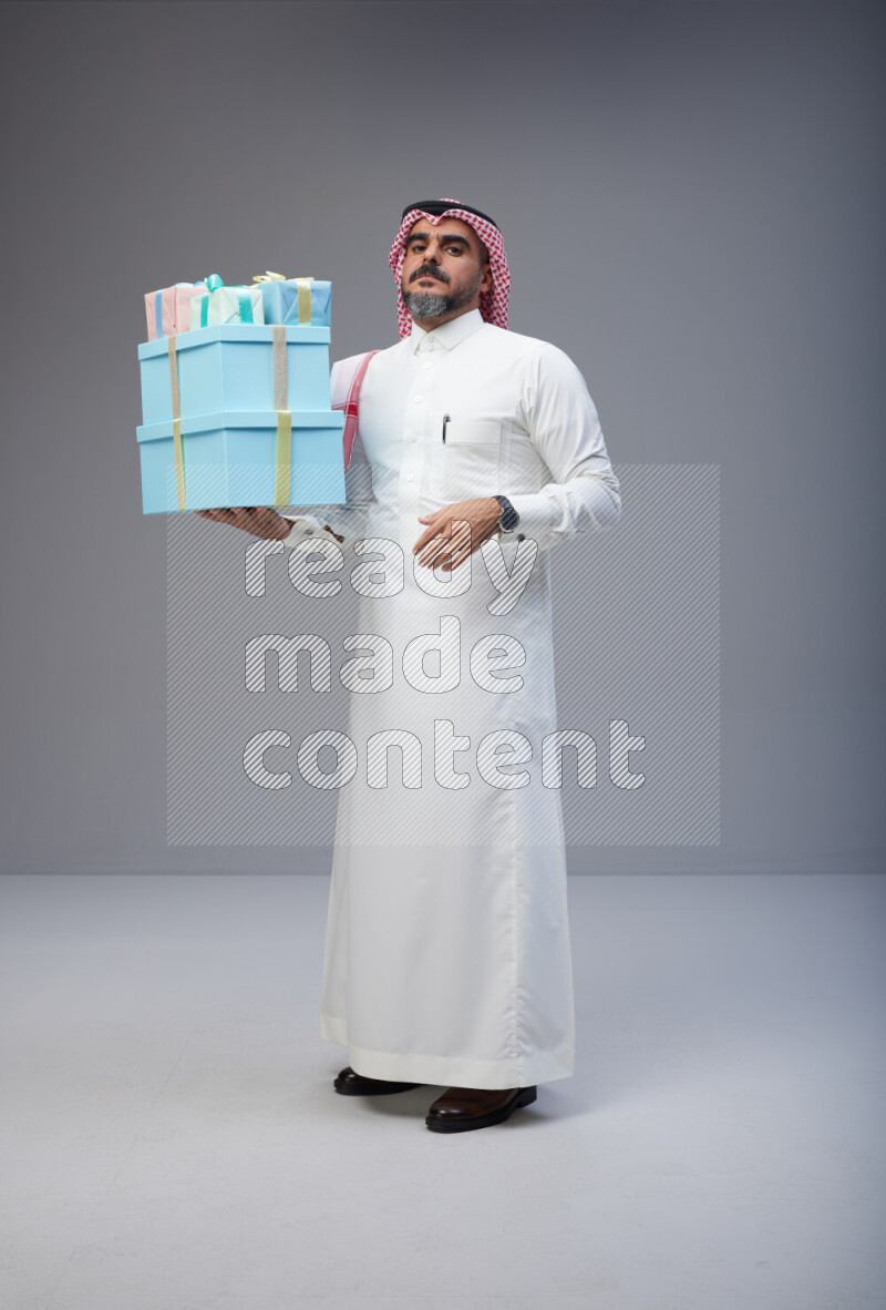 Saudi man Wearing Thob and red Shomag standing holding gift box on Gray background