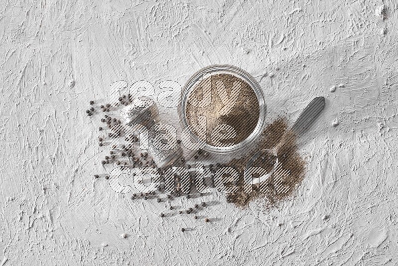 A glass bowl full of black pepper powder with black pepper beads, a turkish metal grinder and a metal spoon on textured white flooring