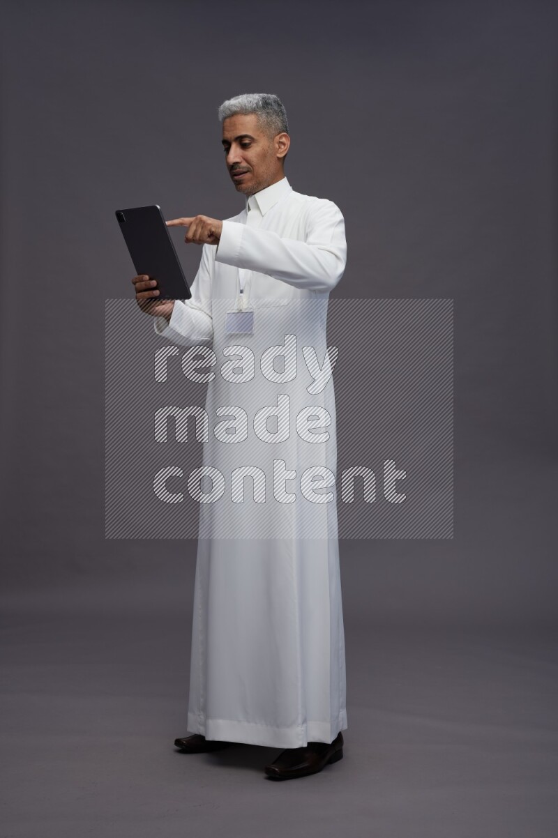 Saudi man wearing thob with neck strap employee badge standing working on tablet on gray background