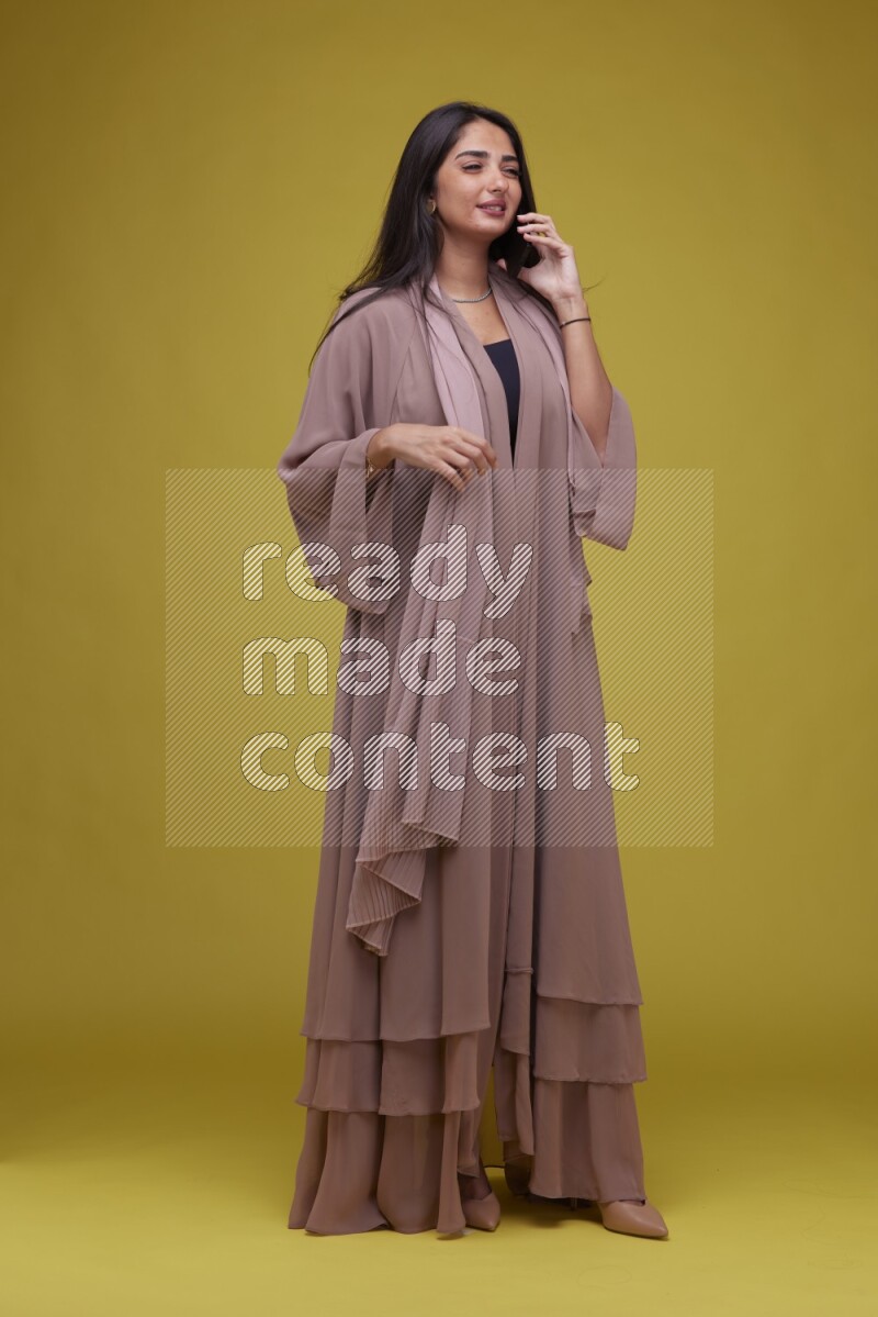 A woman Calling on a Yellow Background wearing Brown Abaya