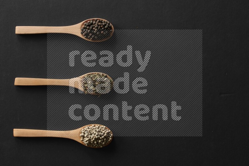 wooden spoons filled with white peppers, cloves, cardamom, salt, black peppers and basil on black flooring