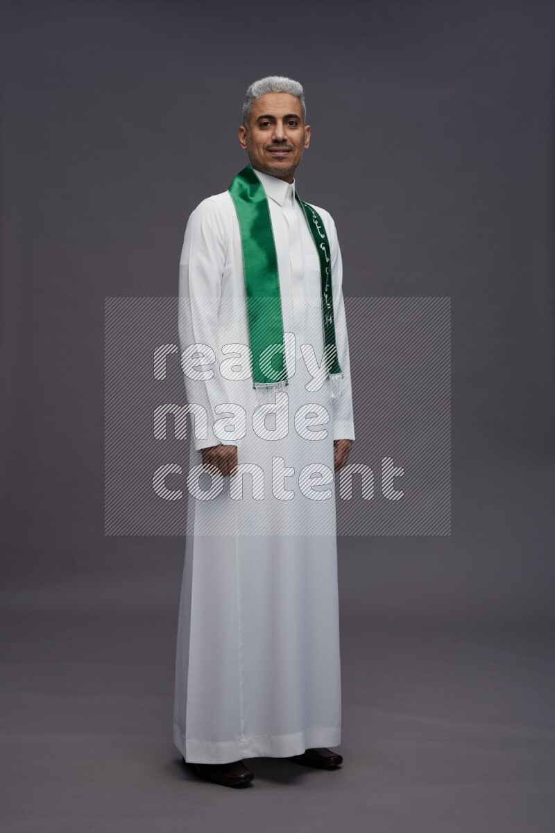 Saudi man wearing thob standing holding Saudi flag on gray background