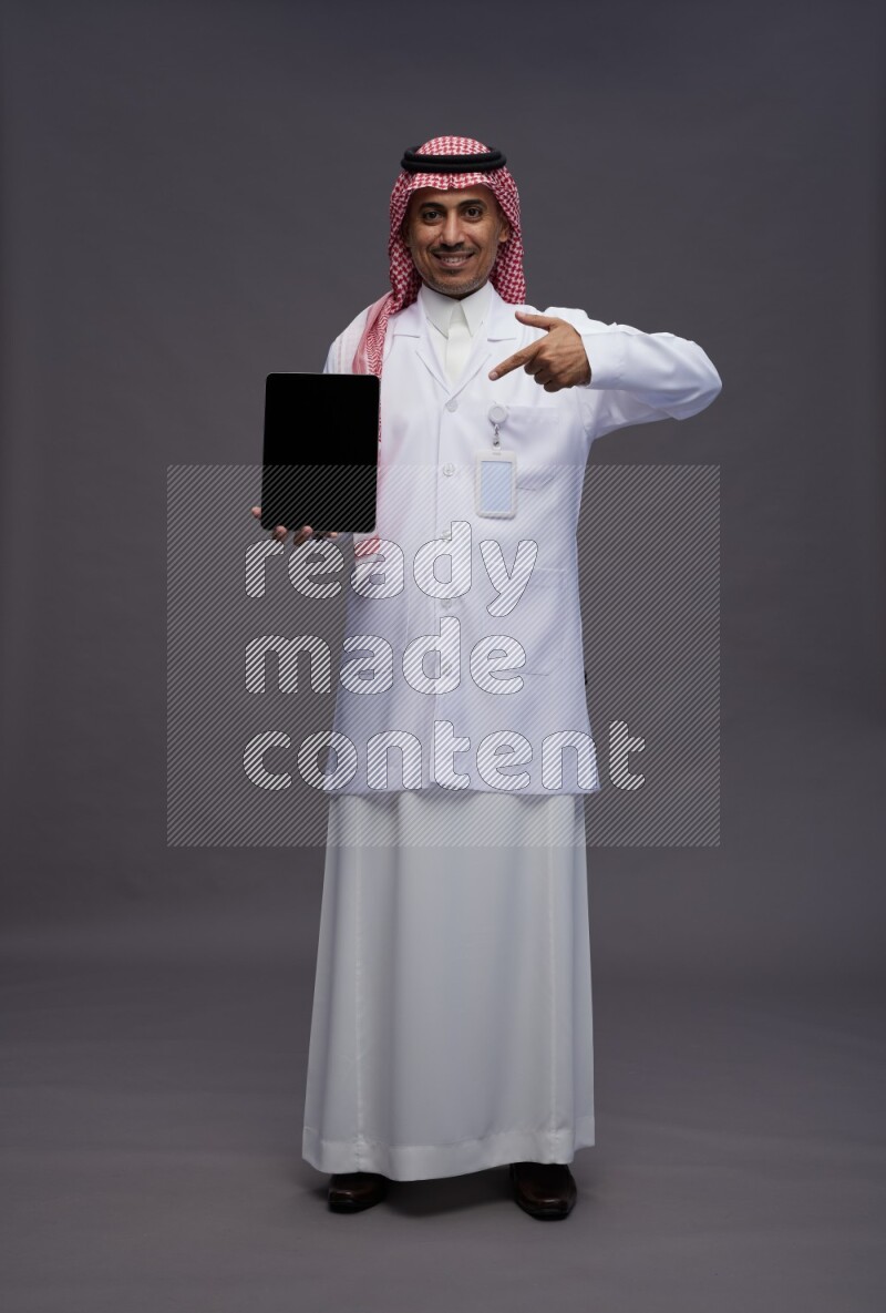Saudi man wearing thob with lab coat and shomag with pocket employee badge standing showing tablet to camera on gray background