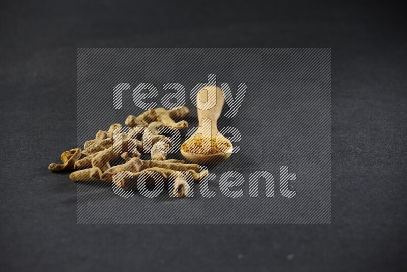 A wooden spoon full of turmeric powder with dried turmeric fingers on black flooring