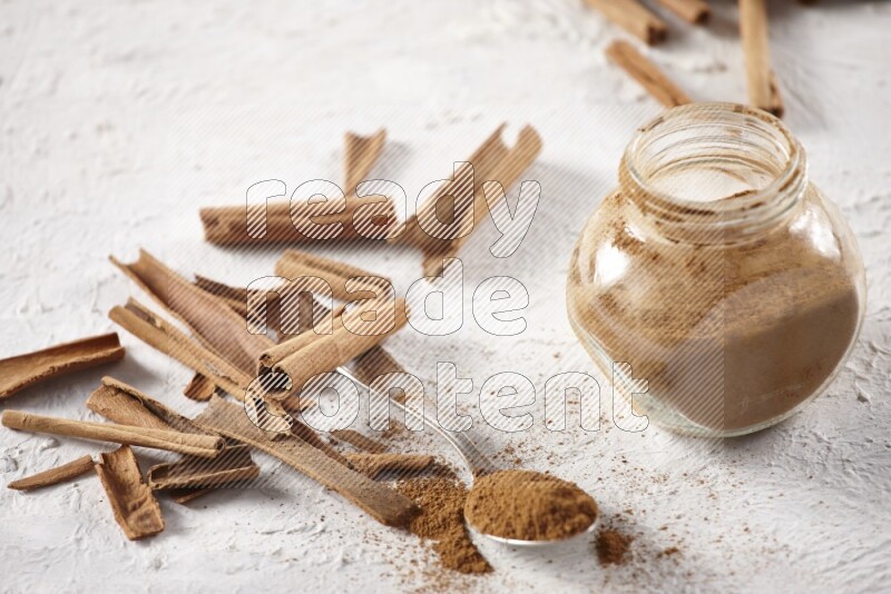Herbal glass jar full cinnamon powder and a metal spoon surrounded by cinnamon sticks on a white background