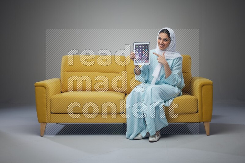A Saudi woman wearing a light blue Abaya and white head scarf sitting on a yellow sofa and showing her tablet's screen while pointing to it eye level on a grey background