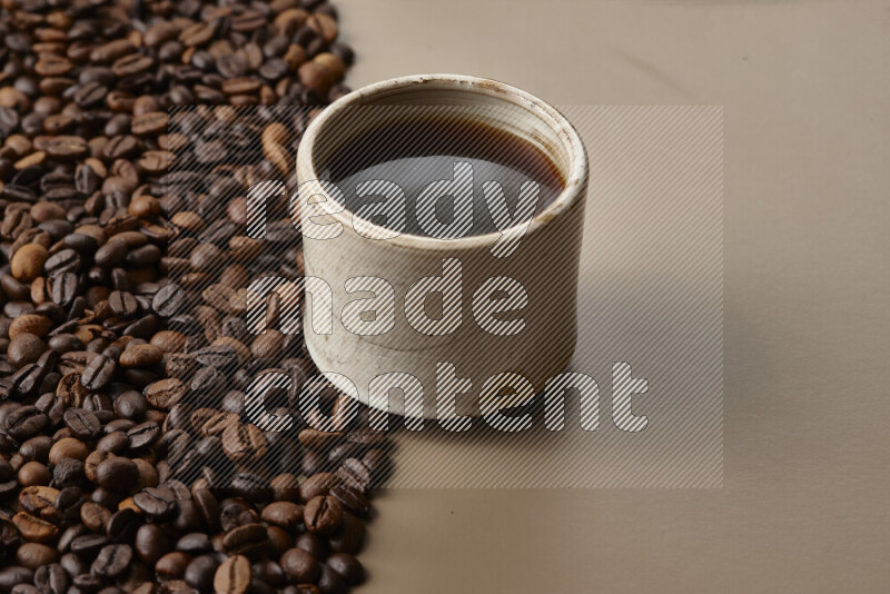 A beige pottery cup of coffee surrounded by roasted coffee beans on beige background