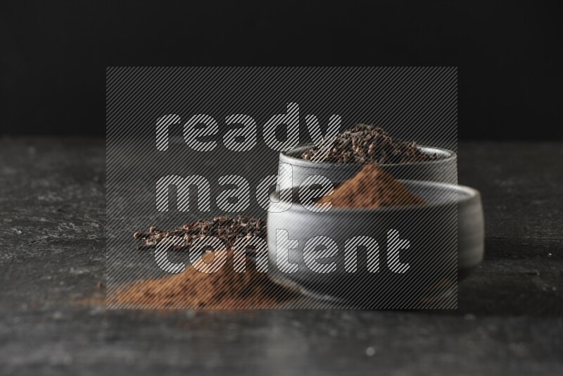 2 Black pottery bowls full of cloves and the other full of cloves powder on textured black flooring