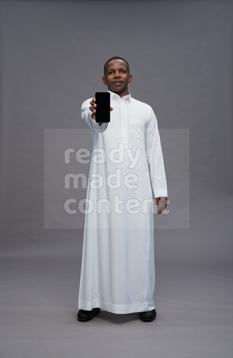 Saudi man wearing thob standing showing phone to camera on gray background