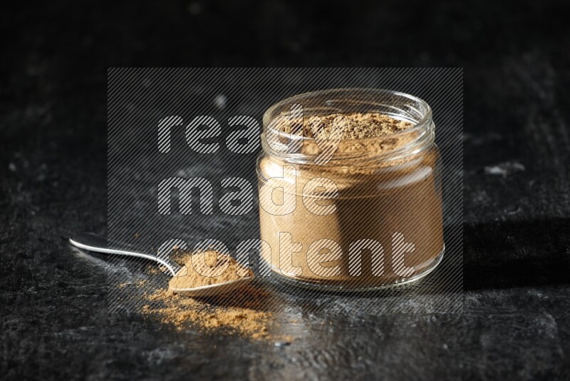 A glass jar and a metal spoon full of allspice powder on a textured black flooring
