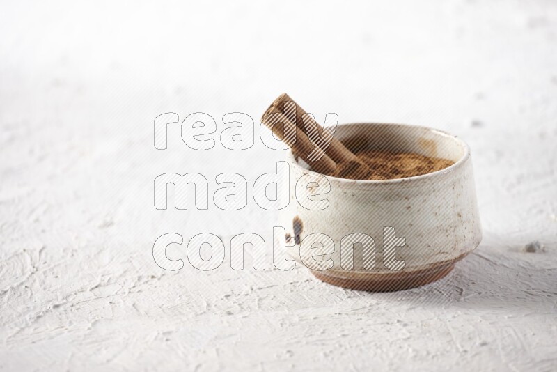 Ceramic beige bowl full of cinnamon powder with a cinnamon stick on a textured white background