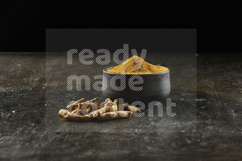 A black pottery bowl full of turmeric powder and dried turmeric whole fingers next of it on textured black flooring