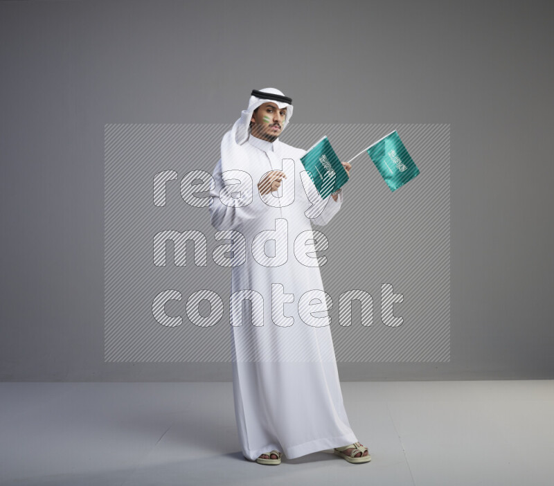 A Saudi man standing wearing thob and white shomag with face painting raising small saudi flag on gray background