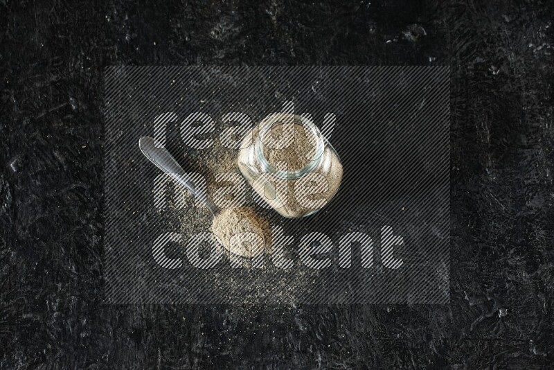 A glass spice jar and metal spoon full of cardamom powder on textured black flooring