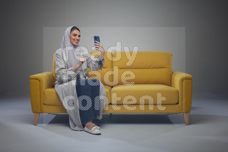 A Saudi woman wearing a light gray Abaya and head scarf sitting on a yellow sofa and making a video call eye level on a grey background